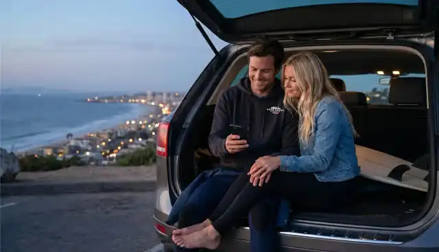 A couple sitting in the back of a car at a Burleigh Heads, Gold Coast lookout at night, wearing a black Sunny souvenir hoodie while using a smartphone to unlock Sunny Link™ VIP access.